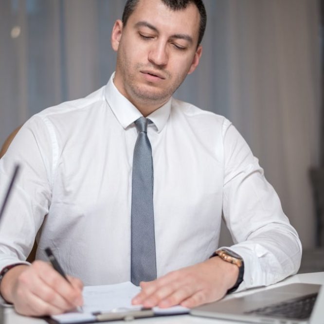 Focused businessman in office writing notes on paper with laptop at a white desk.